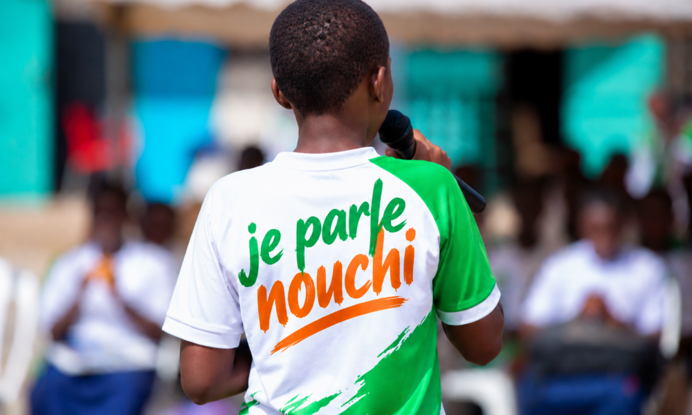 Jeune intervenant vu de dos prenant la parole devant un public, avec l’inscription « je parle nouchi » sur son tee-shirt