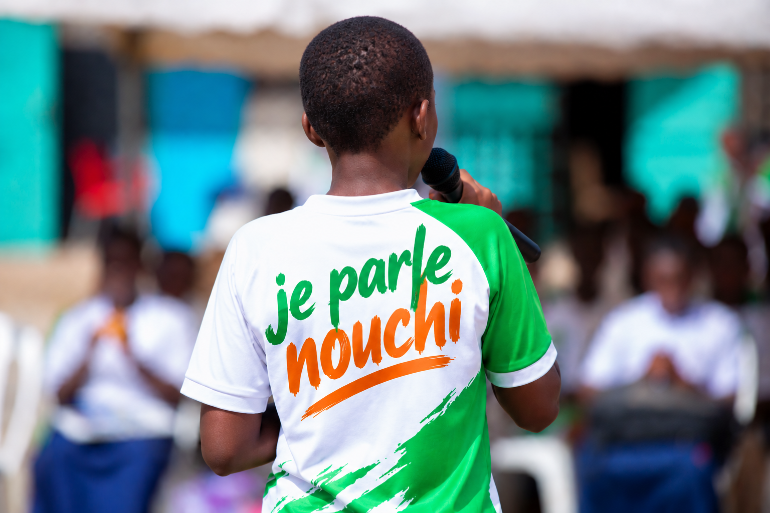 Jeune intervenant vu de dos prenant la parole devant un public, avec l’inscription « je parle nouchi » sur son tee-shirt